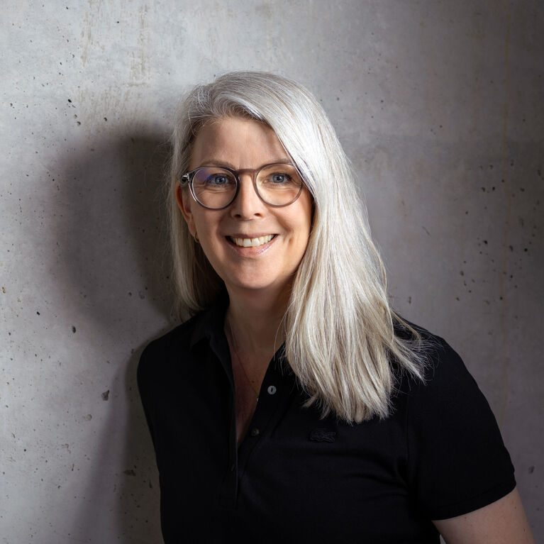 Person mit blondem Haar, Brille und schwarzem Poloshirt vor unscharfem Betonhintergrund (KI-generiert)
