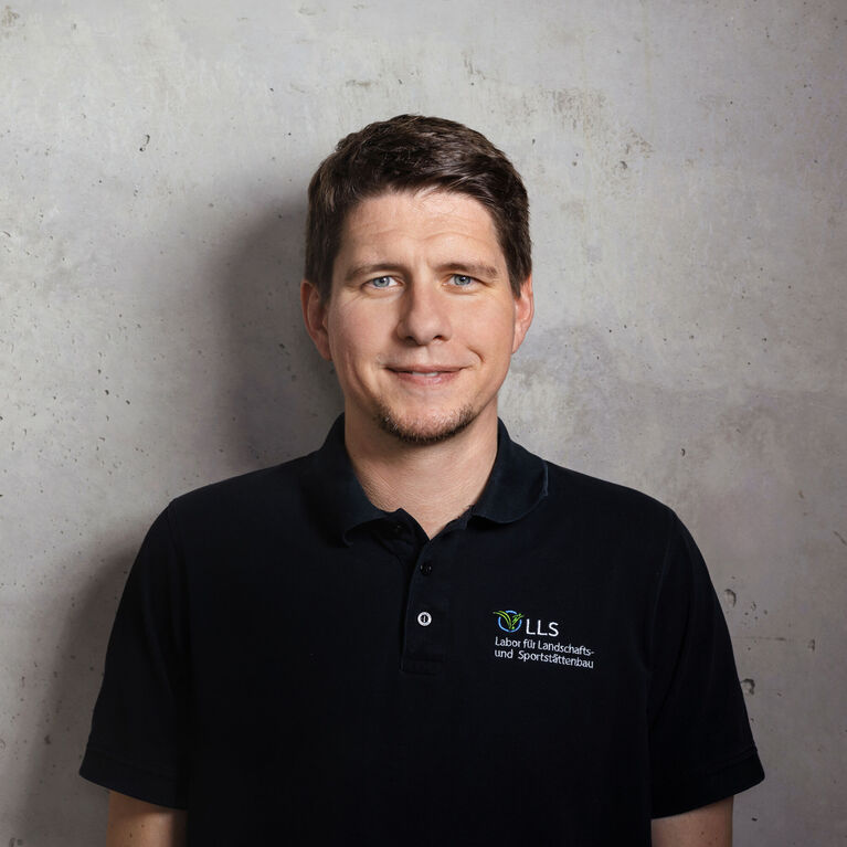 Mann mit kurzen Haaren in schwarzem Poloshirt mit Logo eines Labors für Landschafts- und Sportstättenbau vor unscharfem Hintergrund (KI-generiert)
