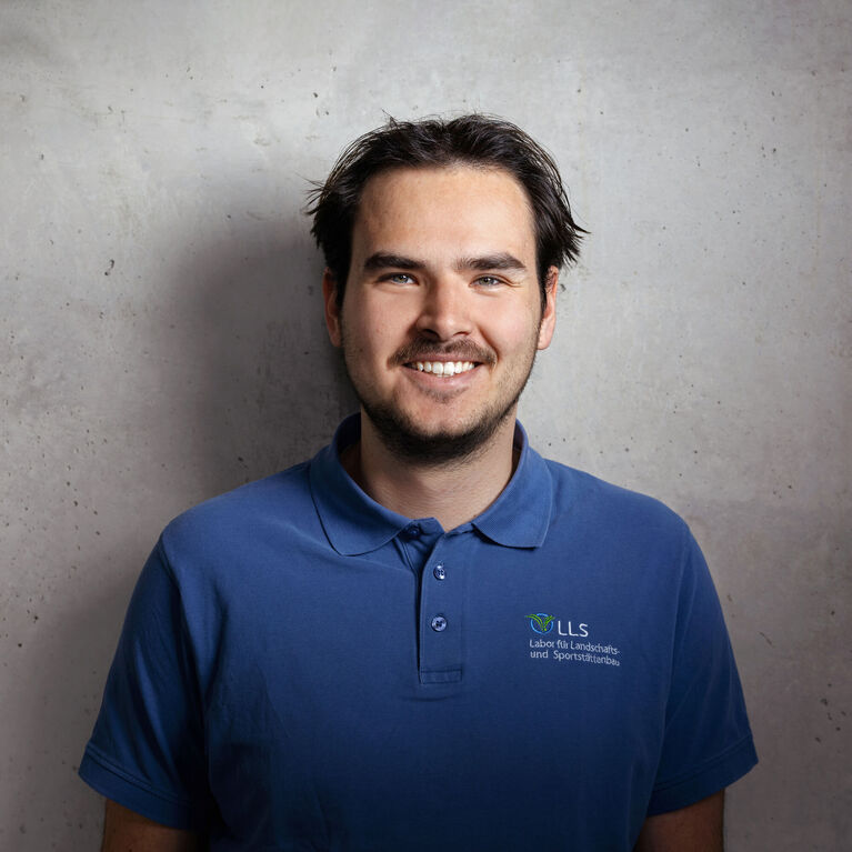Mann mit kurzen dunklen Haaren und blauem Polohemd mit Logo vor strukturierter Betonwand lächelnd (KI-generiert)
