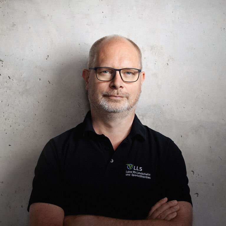 Mann mit Brille und schwarzem Poloshirt mit Logo vor strukturierter Betonwand (KI-generiert)