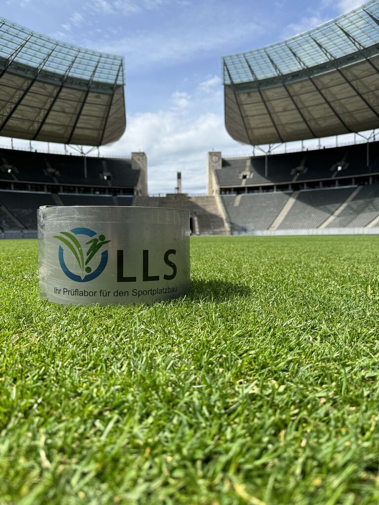 Metallischer Prüfzylinder mit Logo und Text auf einer grünen Rasenfläche in einem Stadion (KI-generiert)