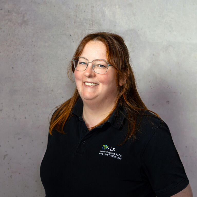 Person mit Brille und dunklem Poloshirt mit Logo eines Labors für Landschafts- und Sportstättenbau vor hellem, unscharfem Hintergrund (KI-generiert)