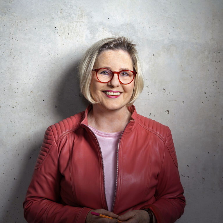 Person mit blondem Kurzhaarschnitt, roter Lederjacke, rosafarbenem Oberteil und runder Brille vor grauem Hintergrund (KI-generiert)