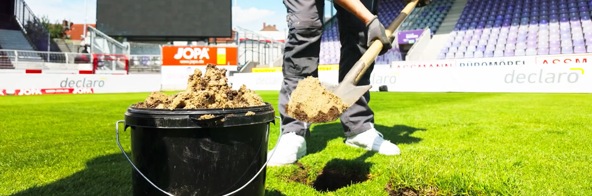 Person mit Spaten füllt Bodenmaterial in einen schwarzen Eimer auf einer Rasenfläche in einem Stadion (KI-generiert)