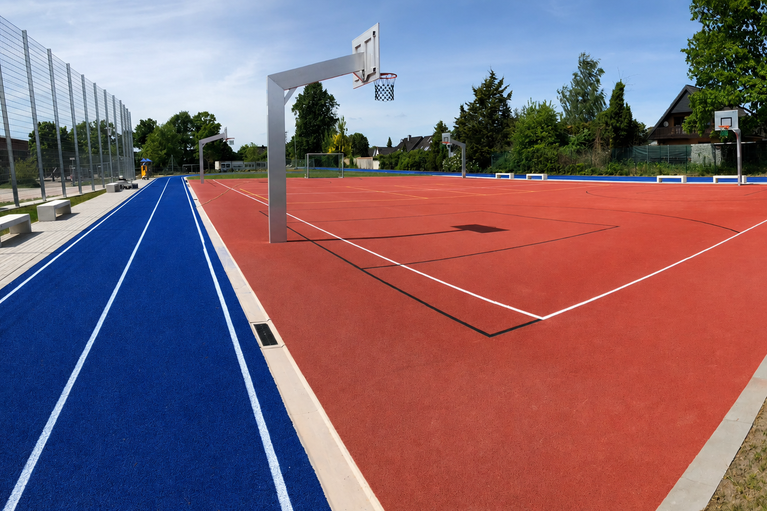 Mehrzwecksportplatz mit rotem Kunststoffbelag, Basketballkorb und Fußballtor auf umgebender Rasenfläche (KI-generiert)