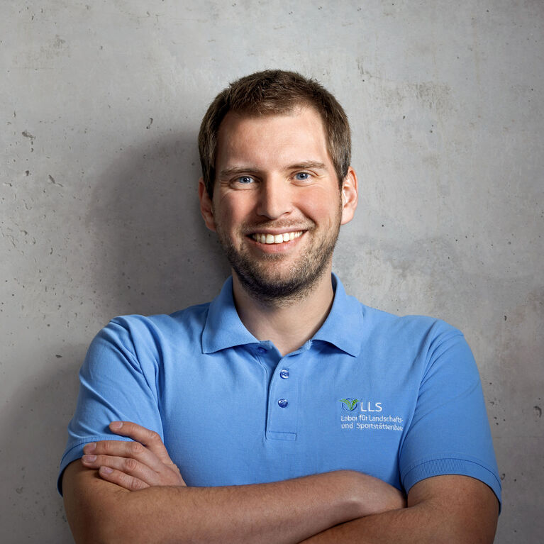 Mann mit kurzen Haaren und blauem Polohemd mit Logo, lächelnd vor unscharfem Betonhintergrund (KI-generiert)