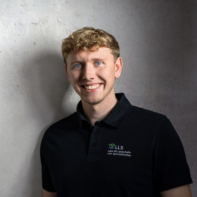 Mann mit lockigem Haar und schwarzem Polohemd mit Logo vor grauem Hintergrund lächelnd (KI-generiert)