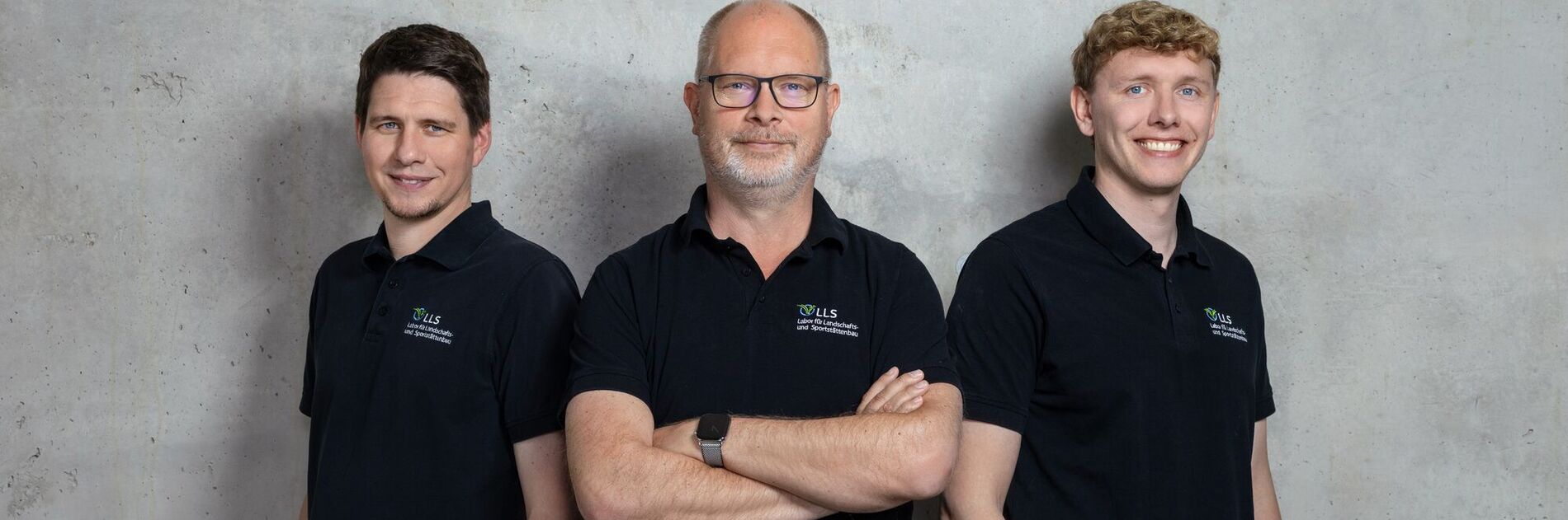 Drei Personen in schwarzen Polo-Shirts mit Logo vor strukturierter Betonwand, lächelnd, Arme verschränkt (KI-generiert)