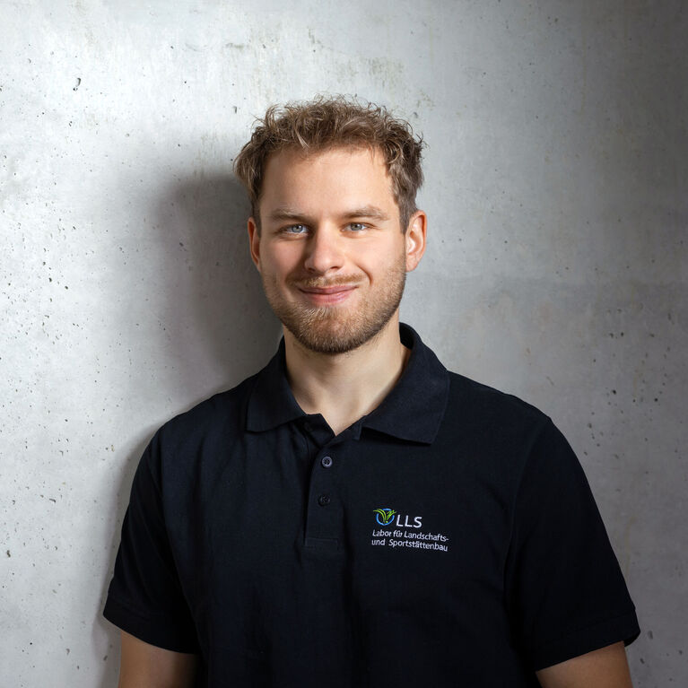 Mann mit kurzen blonden Haaren und Bart in schwarzem Poloshirt mit Logo vor grauer Wand (KI-generiert)