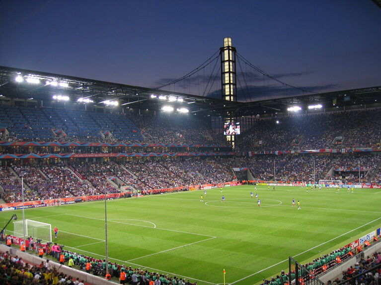 Fußballstadion bei Nacht mit beleuchtetem Naturrasenplatz, vollbesetzten Zuschauerrängen und Flutlichtmasten (KI-generiert)