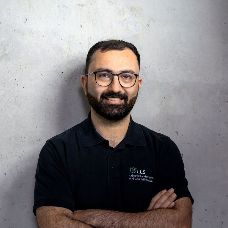 Mann mit Bart und Brille in schwarzem Poloshirt mit Logo eines Labors für Landschafts- und Sportstättenbau vor hellem, unscharfem Hintergrund (KI-generiert)