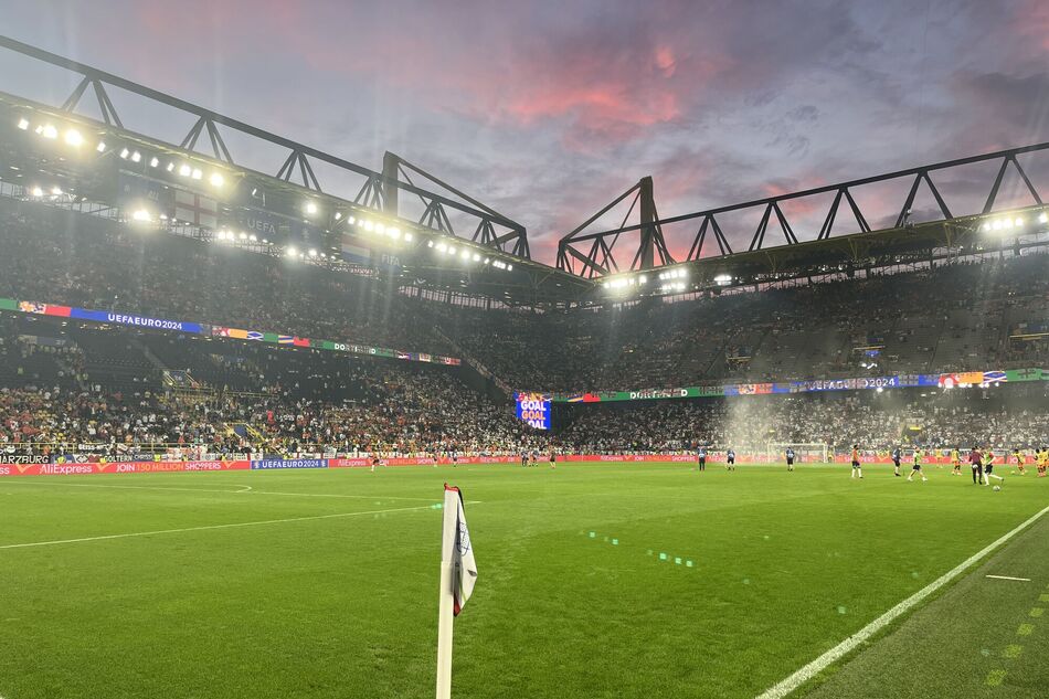 Fußballstadion mit Naturrasenplatz, Flutlichtanlage und gefüllten Zuschauerrängen bei einem Spiel unter künstlicher Beleuchtung (KI-generiert)